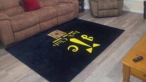 A living room with a brown sofa, a grey armchair, a small table, and a black rug with yellow text. - 3 bedroom with free parking (Schenectady)