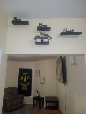Ein Wohnzimmer mit einer Couch, einem kleinen Tisch und einem an der Wand montierten Fernseher. Über der Couch befinden sich dekorative Gegenstände auf Regalen.