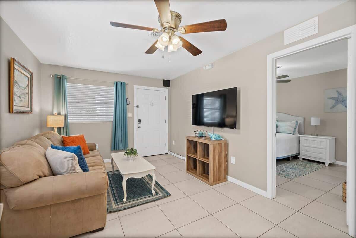 A living room with a ceiling fan, a TV, a sofa, a coffee table, and a door leading to a bedroom.