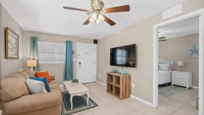 Una sala de estar con ventilador de techo, televisor, sofá, mesa de centro y una puerta que da a un dormitorio.