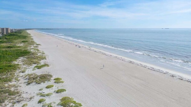 Una playa extensa con olas suaves, cielo despejado y una costa lejana con edificios.
