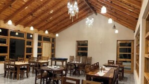 A spacious dining area with wooden tables and chairs, large windows, and a high wooden ceiling with hanging lights. - Bagar Trails  (Kosiyakutoli)