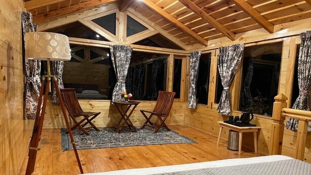 A cozy wooden cabin room with a bed, two chairs, a small table, and a window with curtains. - Bagar Trails  (Kosiyakutoli)
