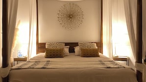 A four-poster canopy bed with a decorative wall piece, two pillows, and a bedside table with a lamp.