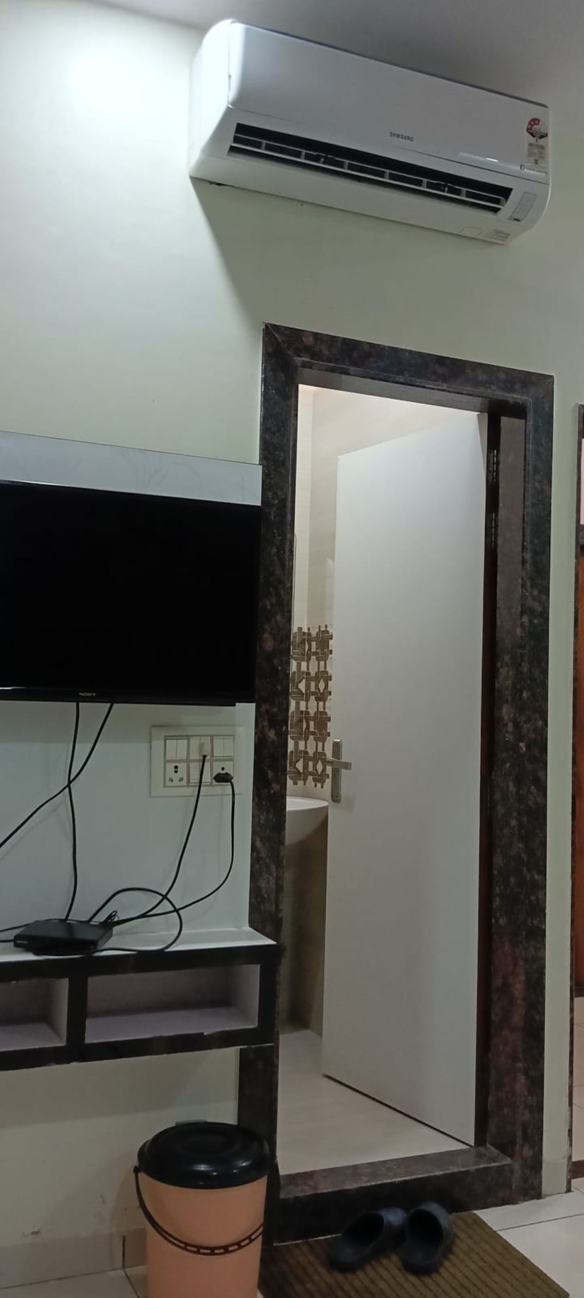 Una habitación con aire acondicionado instalado, una puerta con marco de madera, un estante pequeño con una regleta y un cesto de basura.