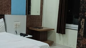 A hotel room with a large bed, a bedside table, a mirror, a small wooden cabinet, and a window with curtains. - Hotel Blue Diamond (Shri Madhopur)