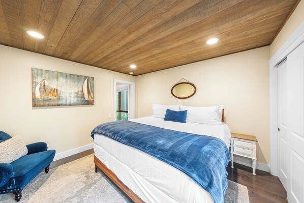 A bedroom with a wooden ceiling, a bed with a blue blanket, a white nightstand, and a blue chair.