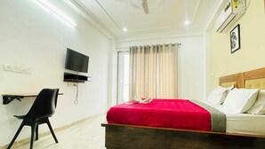 A hotel room with a bed, a desk, a chair, a TV, and a ceiling fan. - OPO MINISTAR M180 (Gurugram)