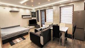 A modern, well-lit room with a bed, sofa, dining table, and a built-in TV.