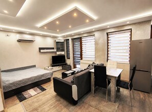 A modern, well-lit room with a bed, sofa, dining table, and a built-in TV. - Elegant Ground-Floor Studio with Covered Terrace and Direct Access to Nature (Пампорово)