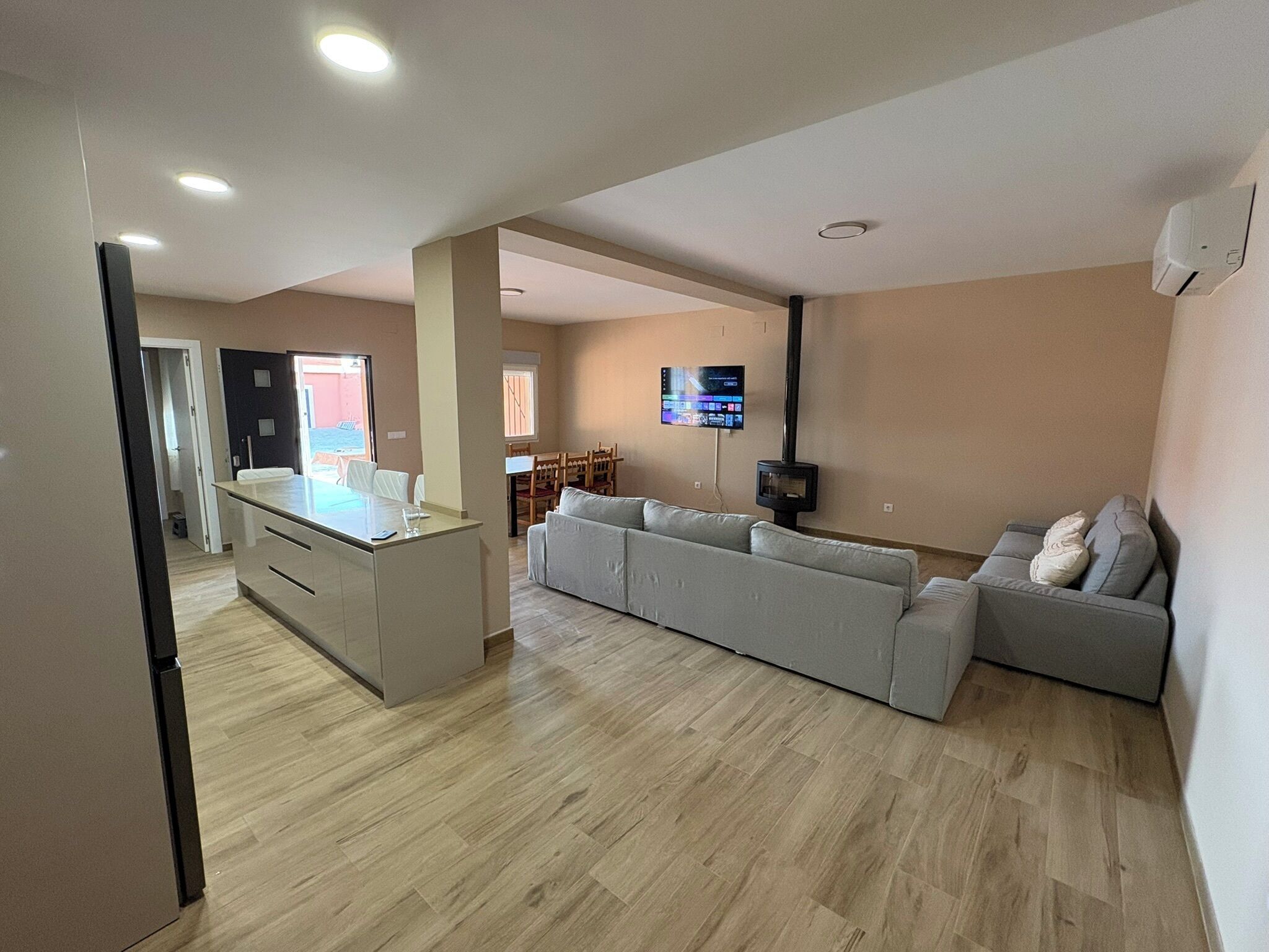 A modern living room with a kitchen area, a large grey sofa, a flat-screen TV mounted on the wall, and a built-in fireplace.