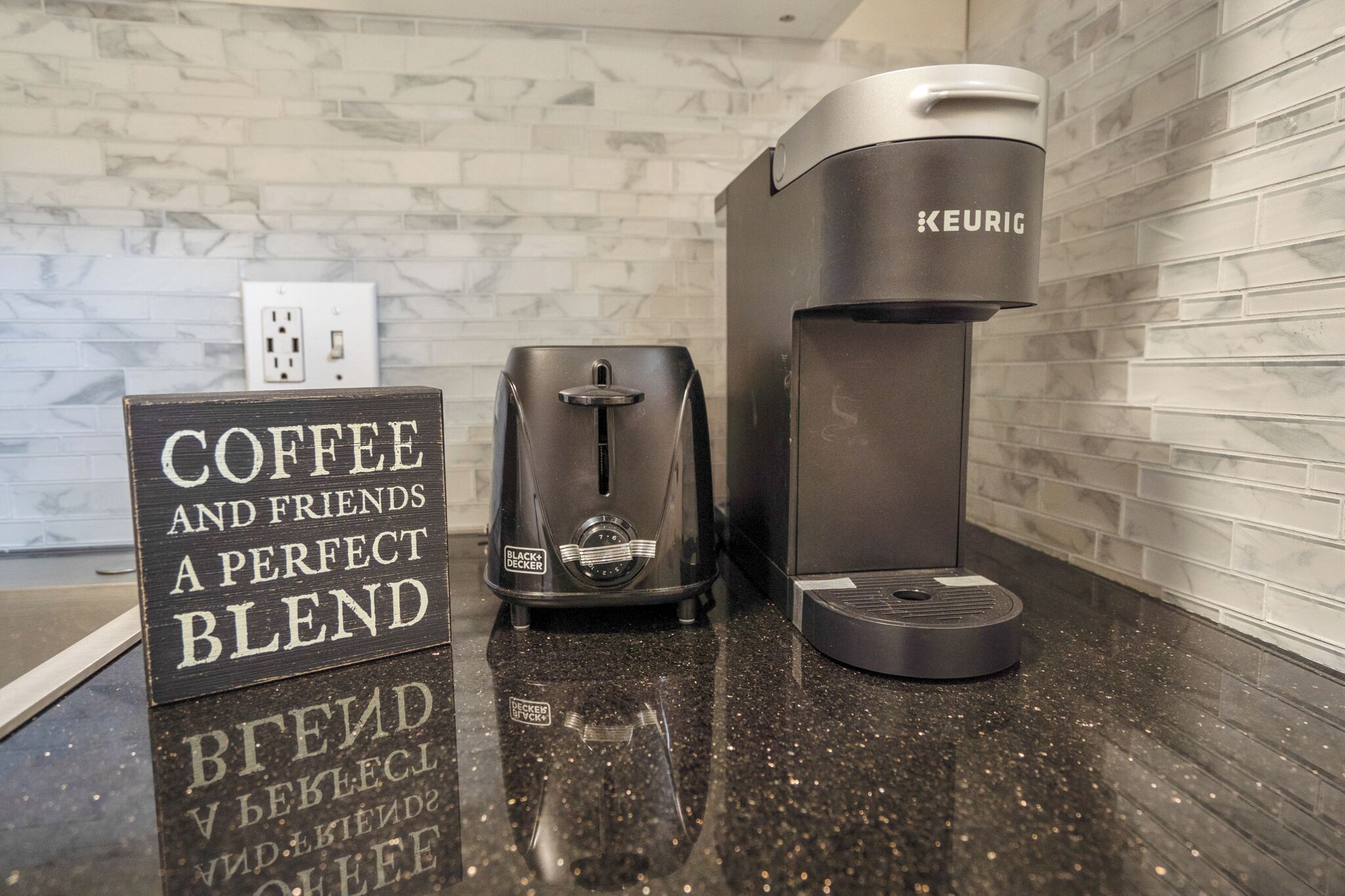 A coffee station with a Keurig and a Black+Decker coffee maker, a sign that reads "COFFEE AND FRIENDS A PERFECT BLEND," and a marble backsplash.