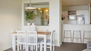 Een moderne keuken met witte kasten, een magnetron en een spoelbak. Er is een eethoek met een houten tafel en witte stoelen.