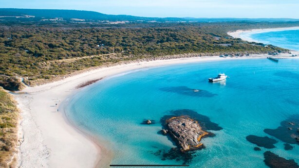 Vue aérienne d’une zone côtière avec une plage de sable, des eaux d’un bleu limpide et un promontoire rocheux au centre.