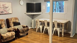 Un salon avec un téléviseur à écran plat fixé au mur, une table à manger avec des chaises et une grande fenêtre.