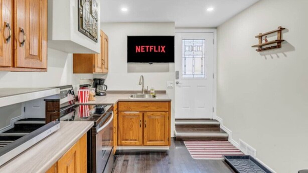 A modern kitchen with wooden cabinets, a built-in oven, and a coffee maker. A flat-screen TV is mounted above the sink, displaying the Netflix logo. - Stylish Zen Stay Mins to Falls | BBQ Area (Niagara Falls)