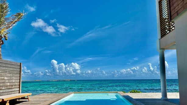 A clear blue pool with a wooden deck, overlooking a turquoise sea and a wooden building. - VIlla Opale — Luxury Living by the Sea (Oyster Pond)