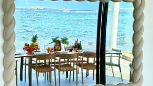 A dining table set with chairs, a fruit bowl, and wine glasses, overlooking a coastal view. - VIlla Opale — Luxury Living by the Sea (Oyster Pond)