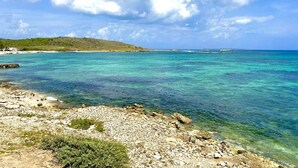 A coastal landscape with clear turquoise waters, rocky shore, and a distant hilly terrain under a partly cloudy sky. - VIlla Opale — Luxury Living by the Sea (Oyster Pond)