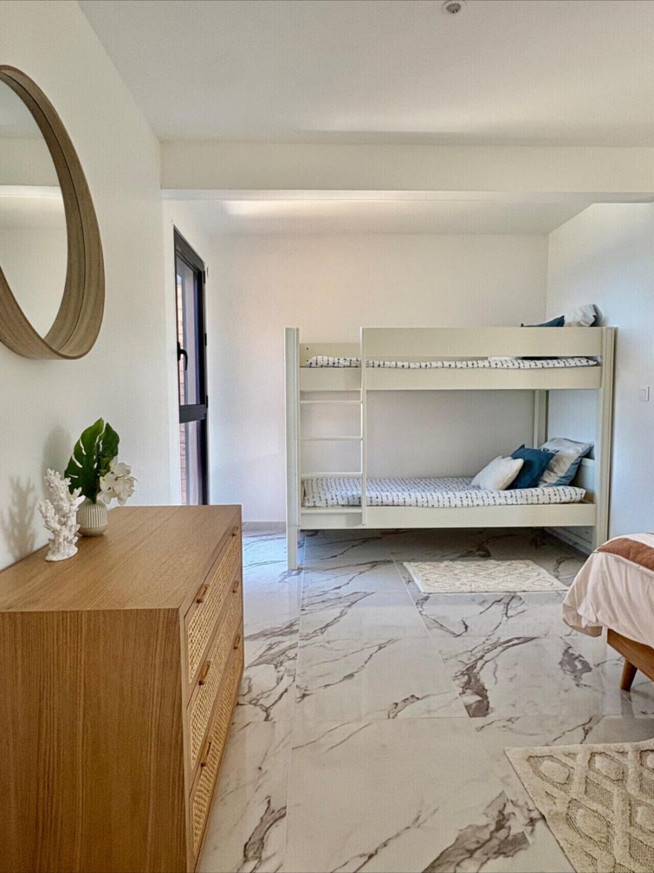 A bunk bed with a white frame and patterned bedding, a wooden dresser with a mirror, and a marble floor.