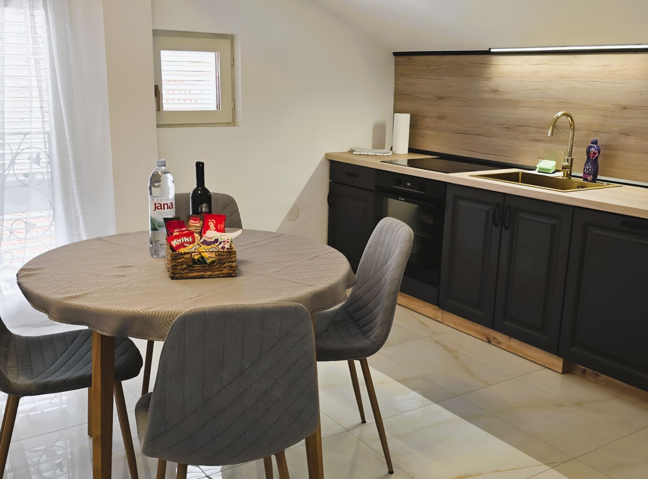 A modern kitchen with a wooden countertop, a sink, and a dishwasher. There is a dining table with chairs and a basket of snacks.