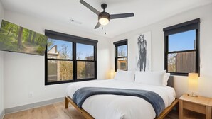 A bedroom with a bed, a ceiling fan, two windows, a nightstand, and a wall-mounted TV.