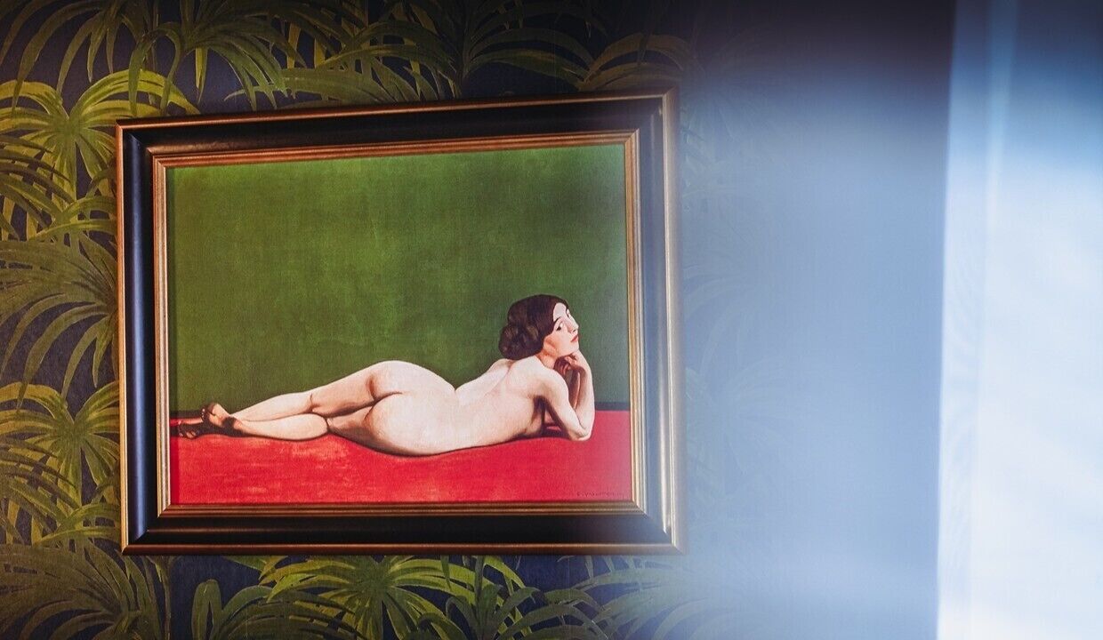 A framed artwork of a reclining nude figure on a red background, set against a wall with a green leafy pattern.