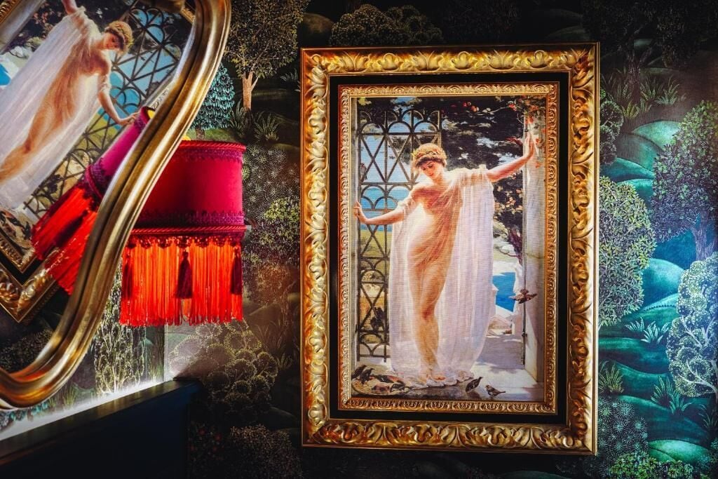 Un cuadro enmarco que muestra a una mujer con vestido blanco, junto a un cojín rojo y flecos decorativos.