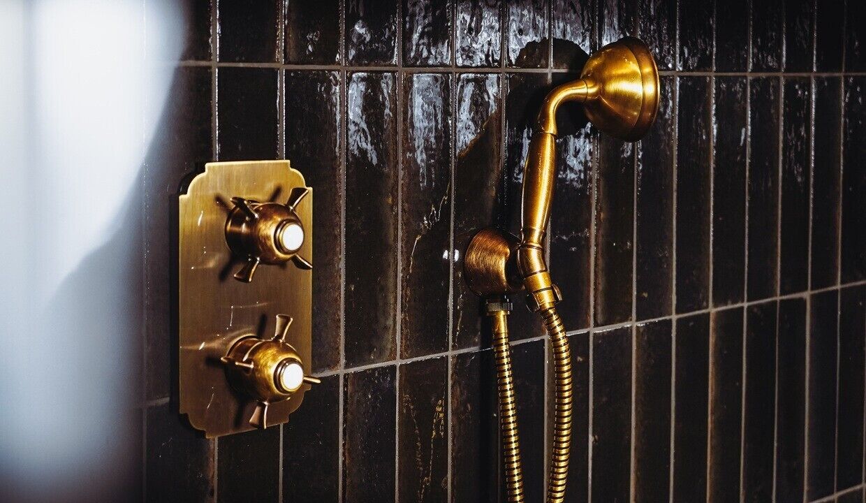Un baño con pared de azulejos negros, un cabezal de ducha de latón y un panel de control con dos perillas.