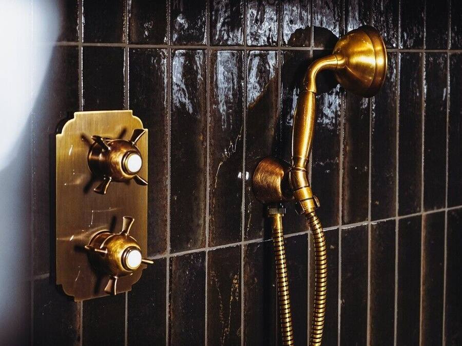Un baño con pared de azulejos negros, un cabezal de ducha de latón y un panel de control con dos perillas.