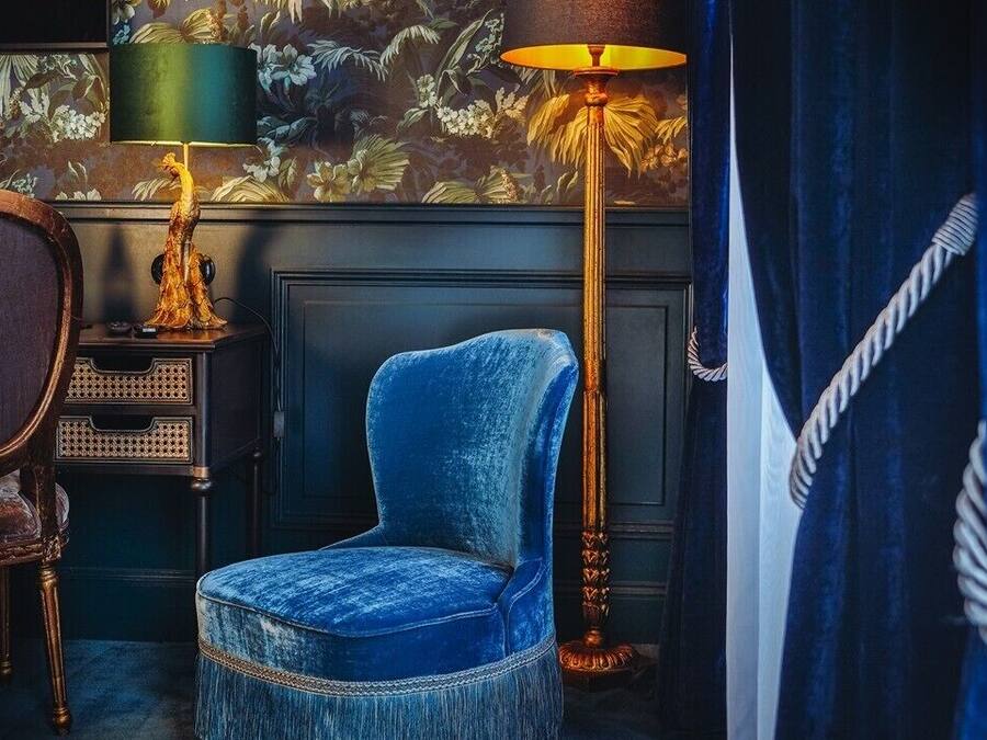 A room with a blue velvet armchair, a floor lamp, a wooden side table, and a patterned wallpaper.