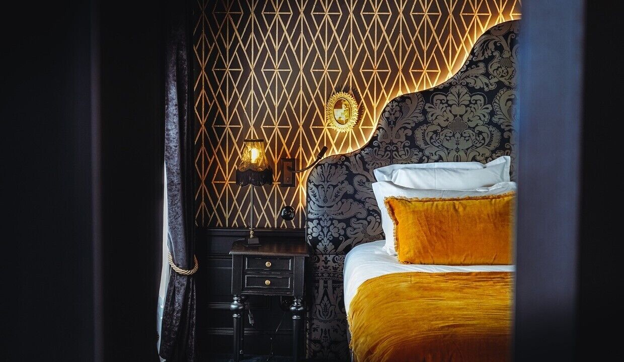 Ein Schlafzimmer mit strukturierter Tapete, einem Bett mit weißer und goldener Bettdecke sowie einem Nachttisch mit Lampe.