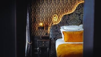 Ein Schlafzimmer mit strukturierter Tapete, einem Bett mit weißer und goldener Bettdecke sowie einem Nachttisch mit Lampe.
