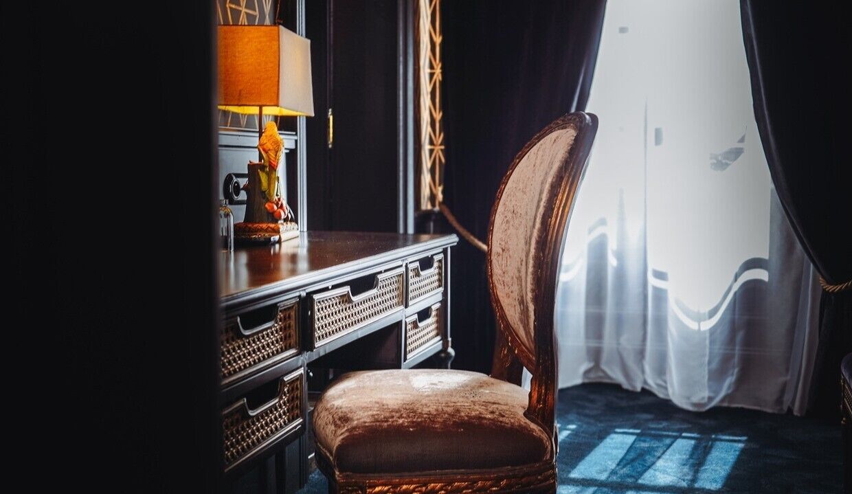 A room with a wooden chair, a desk with drawers, a lamp, and a window with curtains.
