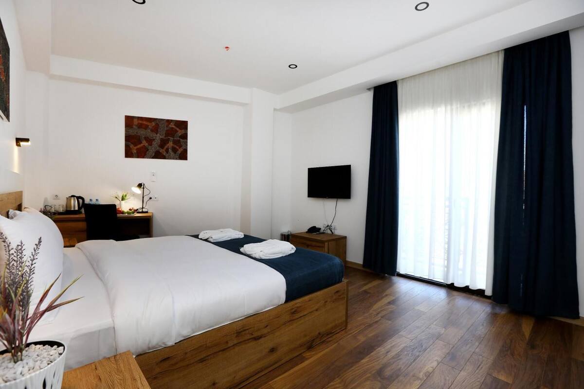 A modern hotel room with a wooden bed, a flat-screen TV, a desk with a lamp, and a large window with curtains.