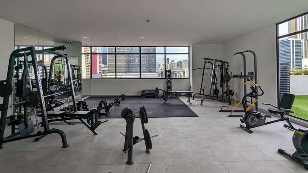 Un gimnasio con diversos equipos de ejercicio, ventanas amplias y vistas a un paisaje urbano.