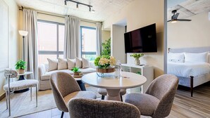 Una habitación de hotel moderna con sofá, dos sillones, una mesa redonda, televisor, cama y un ventanal grande con cortinas.
