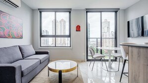Ein modernes Wohnzimmer mit grauem Sofa, einem runden Couchtisch, einem weißen Esstisch mit Stühlen und großen Fenstern mit Blick auf die Stadt.