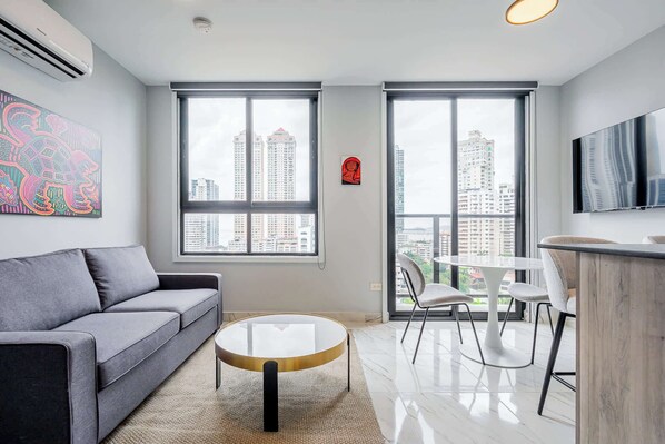 A modern living room with a grey sofa, a round coffee table, a white dining table with chairs, and large windows offering a city view.