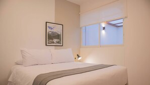A bedroom with a bed, a framed picture on the wall, a window with blinds, and a bedside lamp.