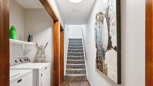 A narrow hallway with a wooden floor, a washing machine, and a dryer on the left, and a staircase leading to an upper floor.
