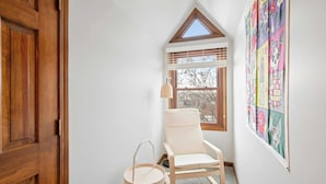A corner room with a wooden door, a white chair, a small round table, a window with blinds, and a colorful wall hanging.