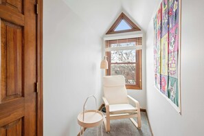 Ein Eckzimmer mit Holztür, einem weißen Stuhl, einem kleinen runden Tisch, einem Fenster mit Jalousien und einem farbenfrohen Wandbehang.