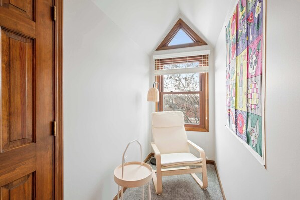 Ein Eckzimmer mit Holztür, einem weißen Stuhl, einem kleinen runden Tisch, einem Fenster mit Jalousien und einem farbenfrohen Wandbehang.