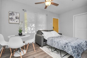 Quarto moderno com cama, sofá cinza, mesinha com cadeiras e ventoinha no teto.