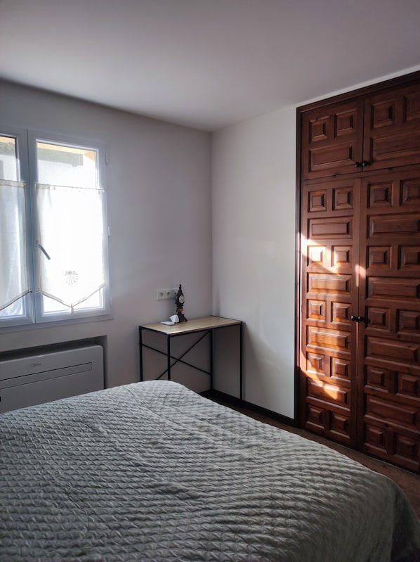 A bedroom with a bed, a small table, a wooden wardrobe, and a window.