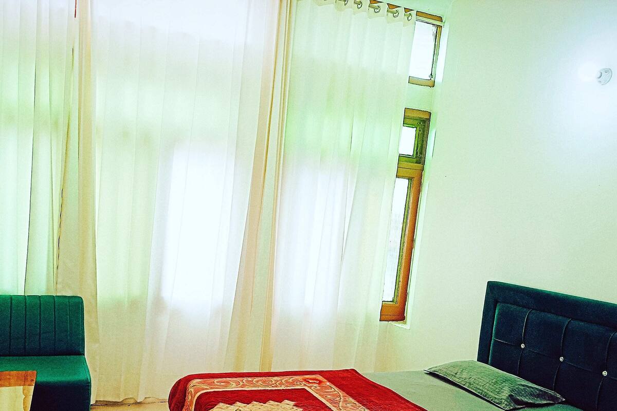 A bedroom with a bed, a green armchair, a small table, and a window with curtains.