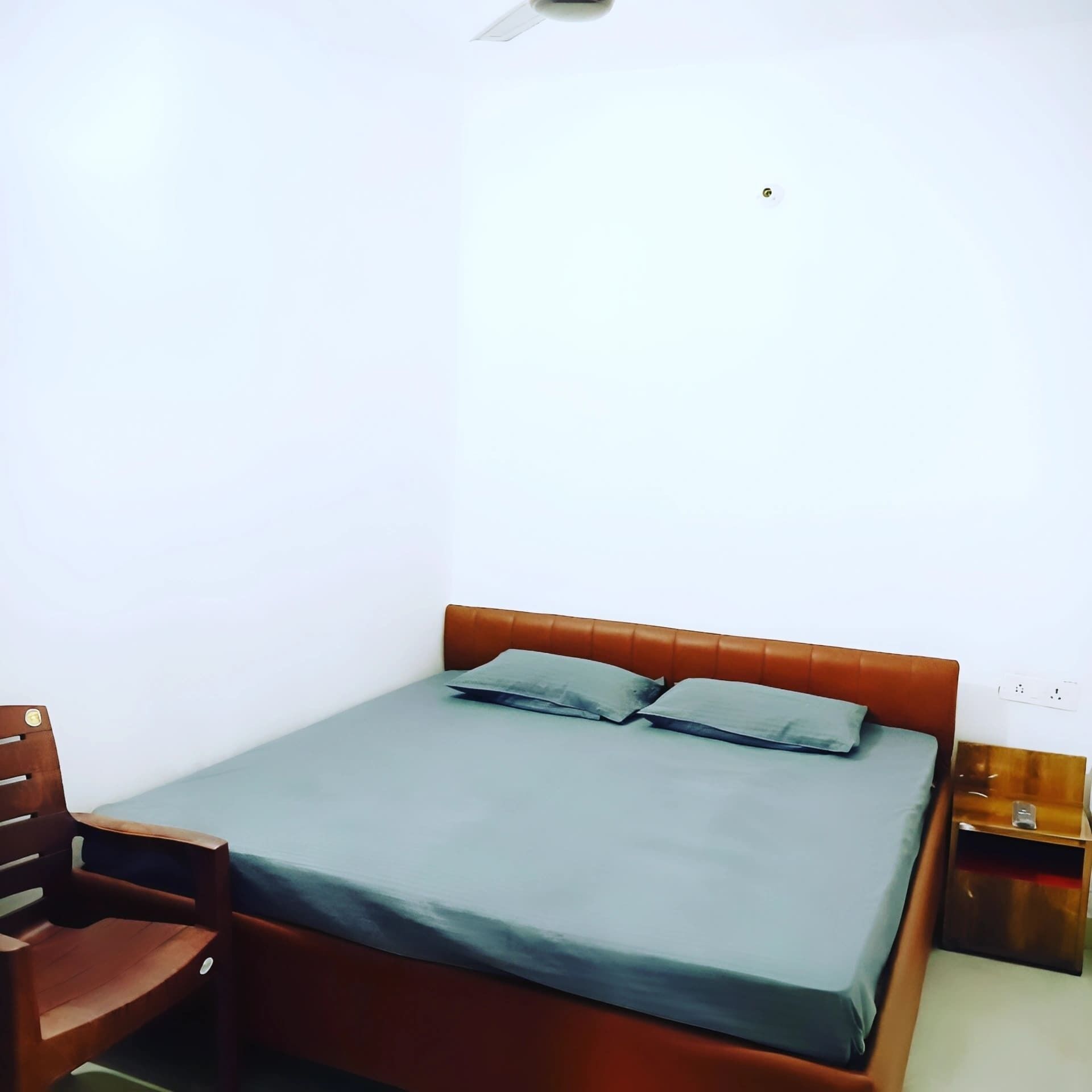 A single bed with a grey mattress and two pillows, a wooden chair, a bedside table with a lamp, and a power outlet on the wall.