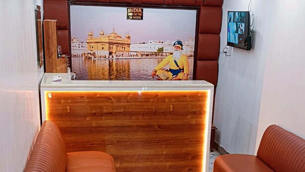 Zona de recepción con mostrador de madera, dos sofás marrones y un televisor mostrando una escena con una persona vestida con traje tradicional.