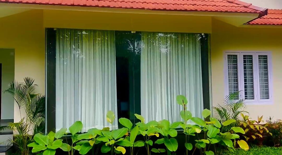 Un edificio amarillo con techo rojo, ventanas grandes con cortinas blancas y plantas verdes en primer plano.